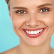 Portrait d'une femme souriante aux yeux verts et aux dents blanches parfaites, posant devant un fond bleu clair.