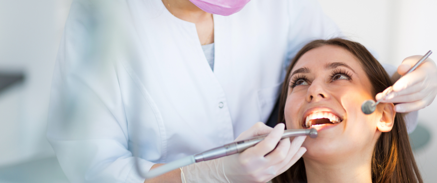 Un dentiste en blouse blanche et masque rose traite une patiente souriante allongée sur une chaise dentaire, utilisant des instruments dentaires pour un examen ou un traitement.