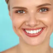 Portrait d'une femme souriante aux yeux verts et aux dents blanches parfaites, posant devant un fond bleu clair.